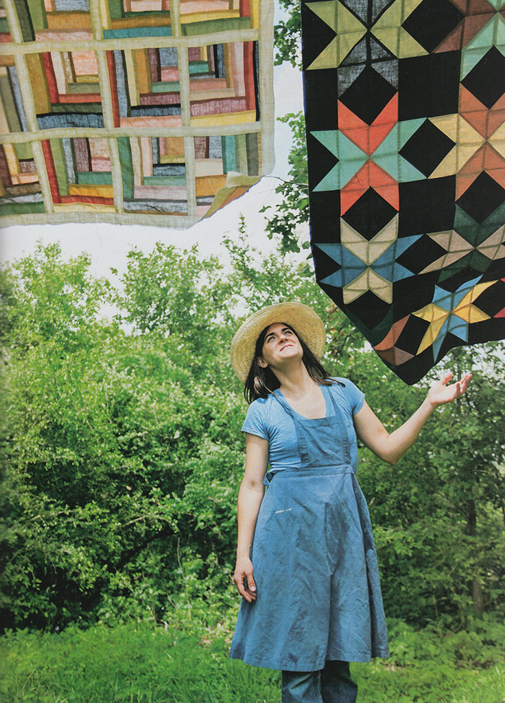lady wearing a straw hat and blue apron looking up at 2 colorful quilts on clothesline 